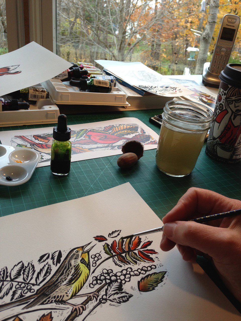 Claudia McGehee's desk filled with scraperboard artwork, with a view out to the woods